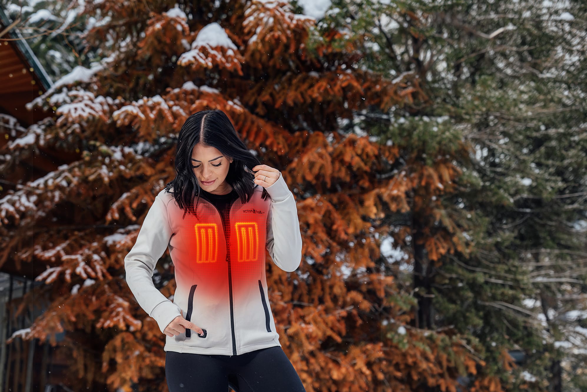 Woman in white jacket with glowing red heating panels stands in snowy forest, adjusting hair and pocket.